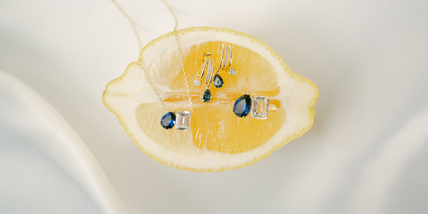 Gold jewelry set featuring a necklace, earrings, and a ring, all set with pear-shaped blue and white sapphire gemstones, displayed creatively on a slice of bright yellow lemon.