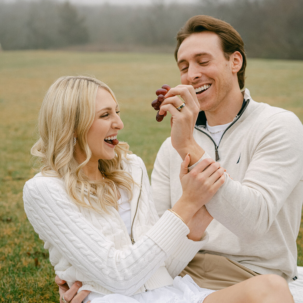 Joyful and playful portrait of a couple laughing while feeding each other red grapes outdoors. The woman wears a white cable-knit jacket and skirt, and the man wears a light sweater, sitting on a grassy field on a misty day.
