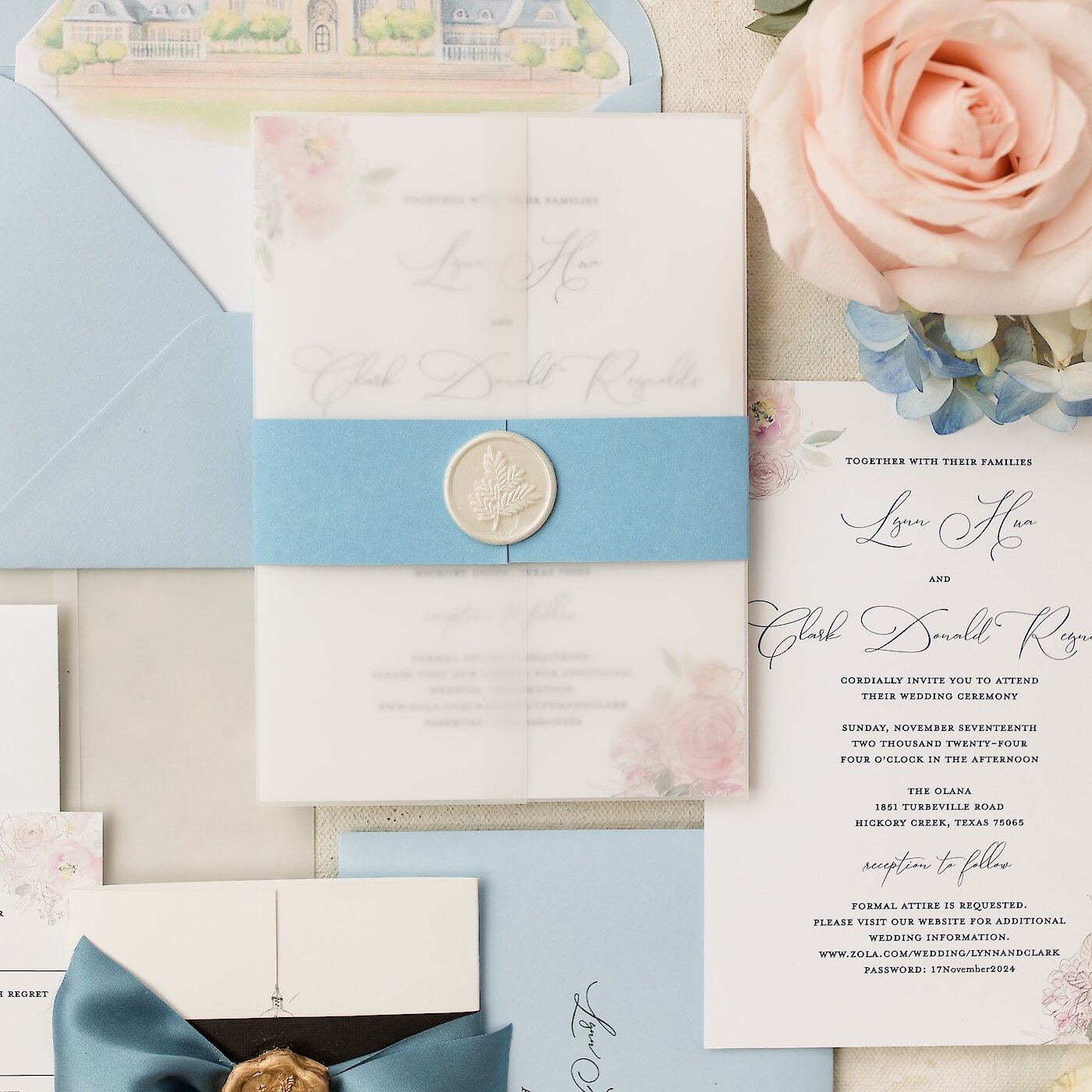 A wedding invitation suite flat lay featuring dusty blue accents, vellum paper, a floral wax seal, and a blush pink rose.
