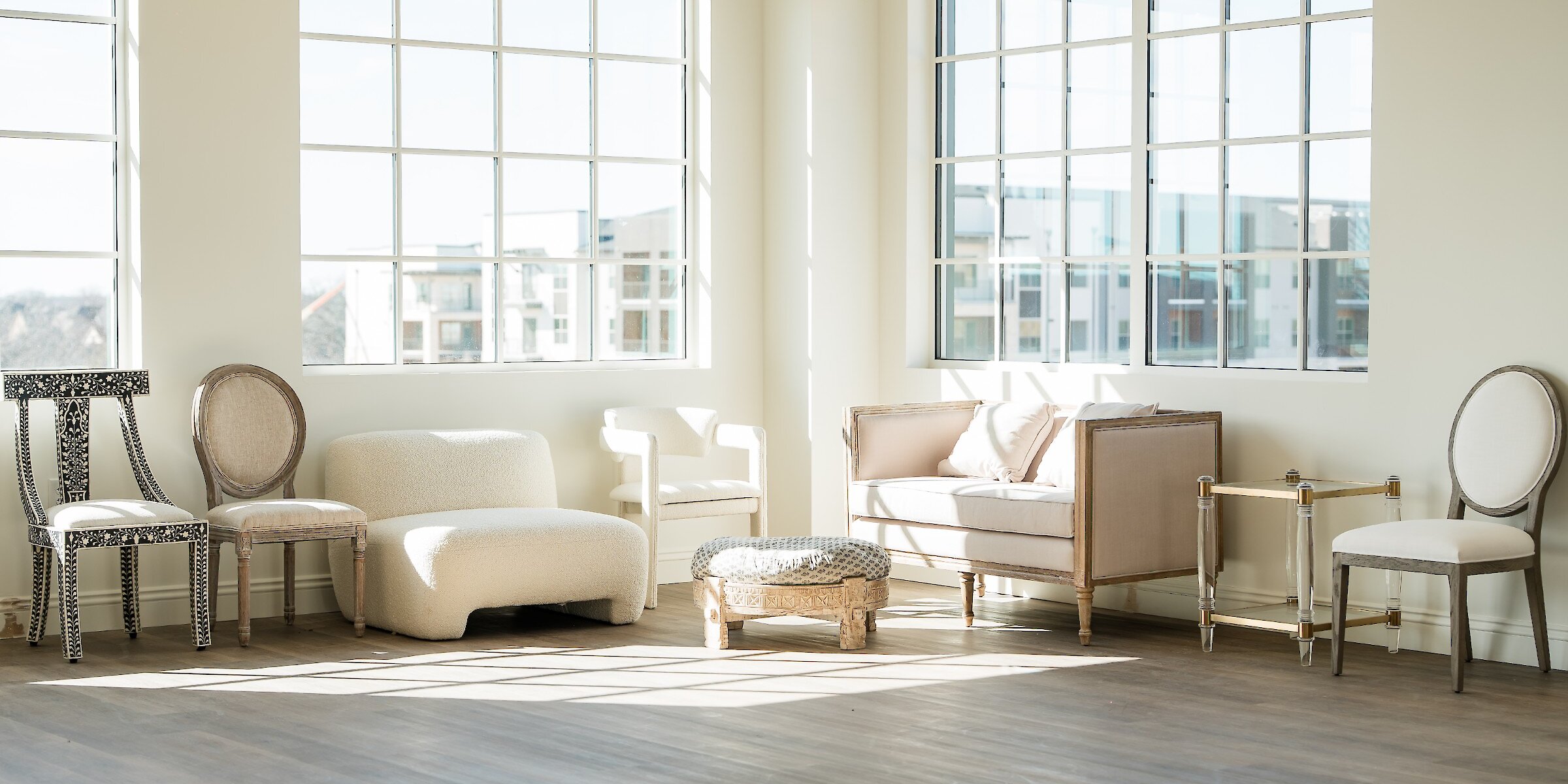 Sunlit photo studio with hardwood floors featuring an eclectic collection of neutral-toned lounge furniture, including chairs and a small couch.