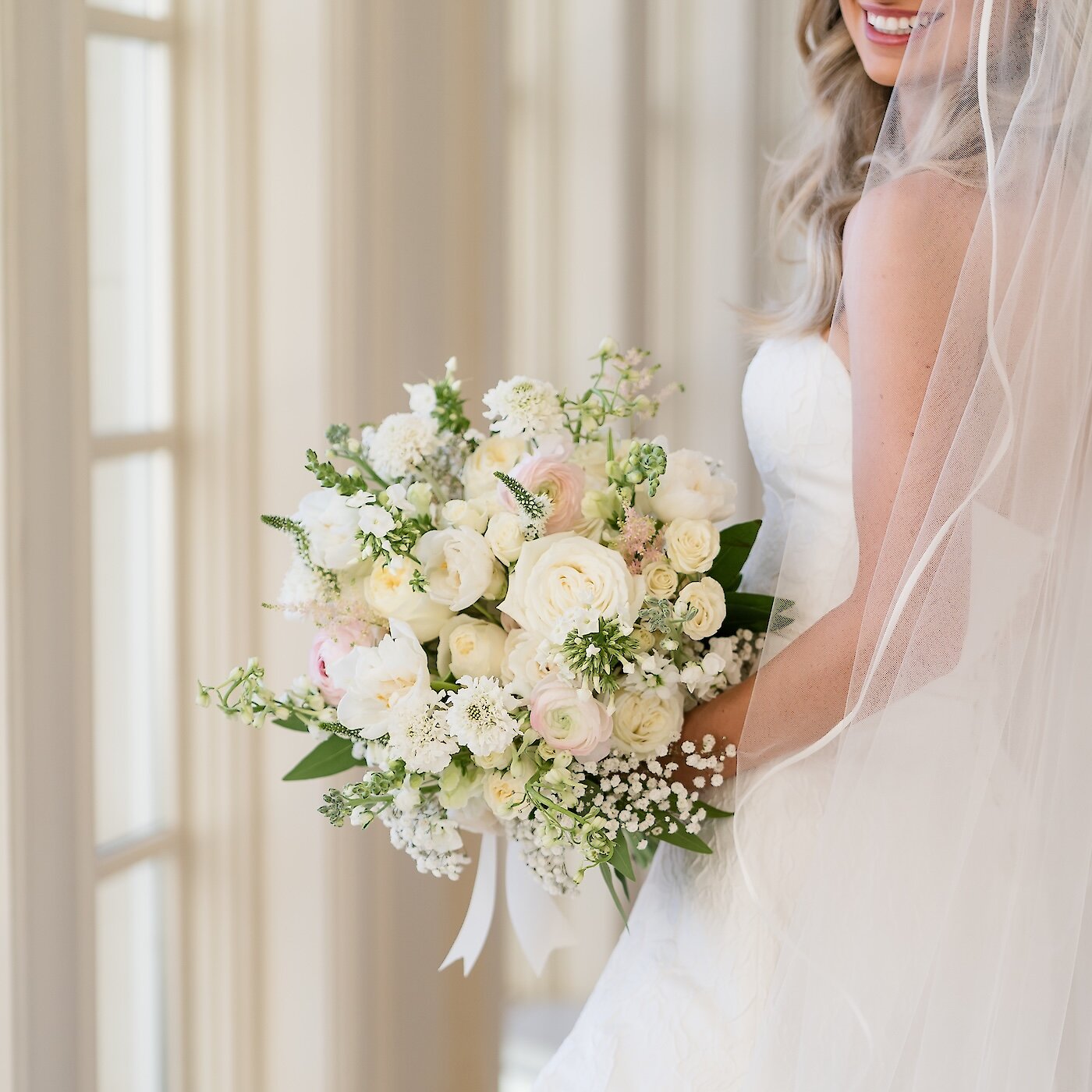 The bride holds a lush, textured bouquet of white and pale pink roses, ranunculus, and baby's breath near a window.