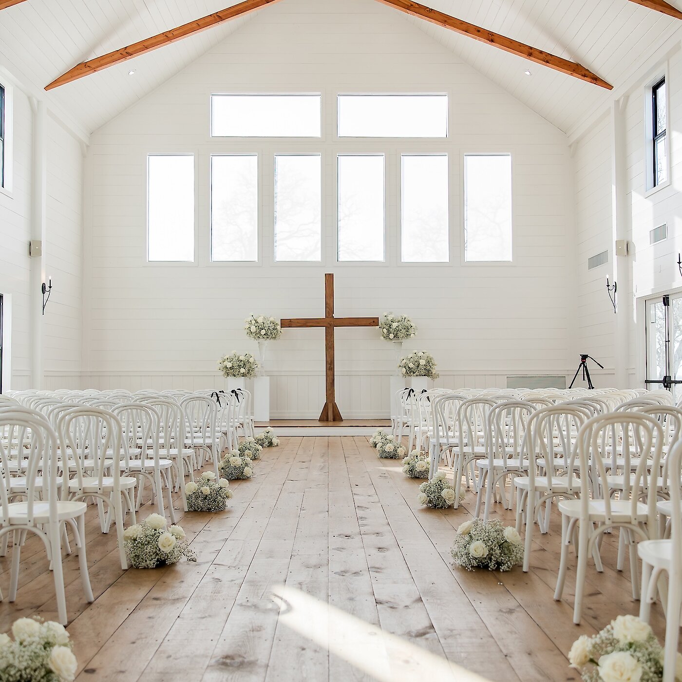 Bright, minimalist white wedding chapel interior with a vaulted ceiling, exposed wooden beams, and white horizontal plank walls. The altar features a large wooden cross, flanked by white floral arrangements, with the aisle lined with white chairs and small white and baby's breath floral mounds.