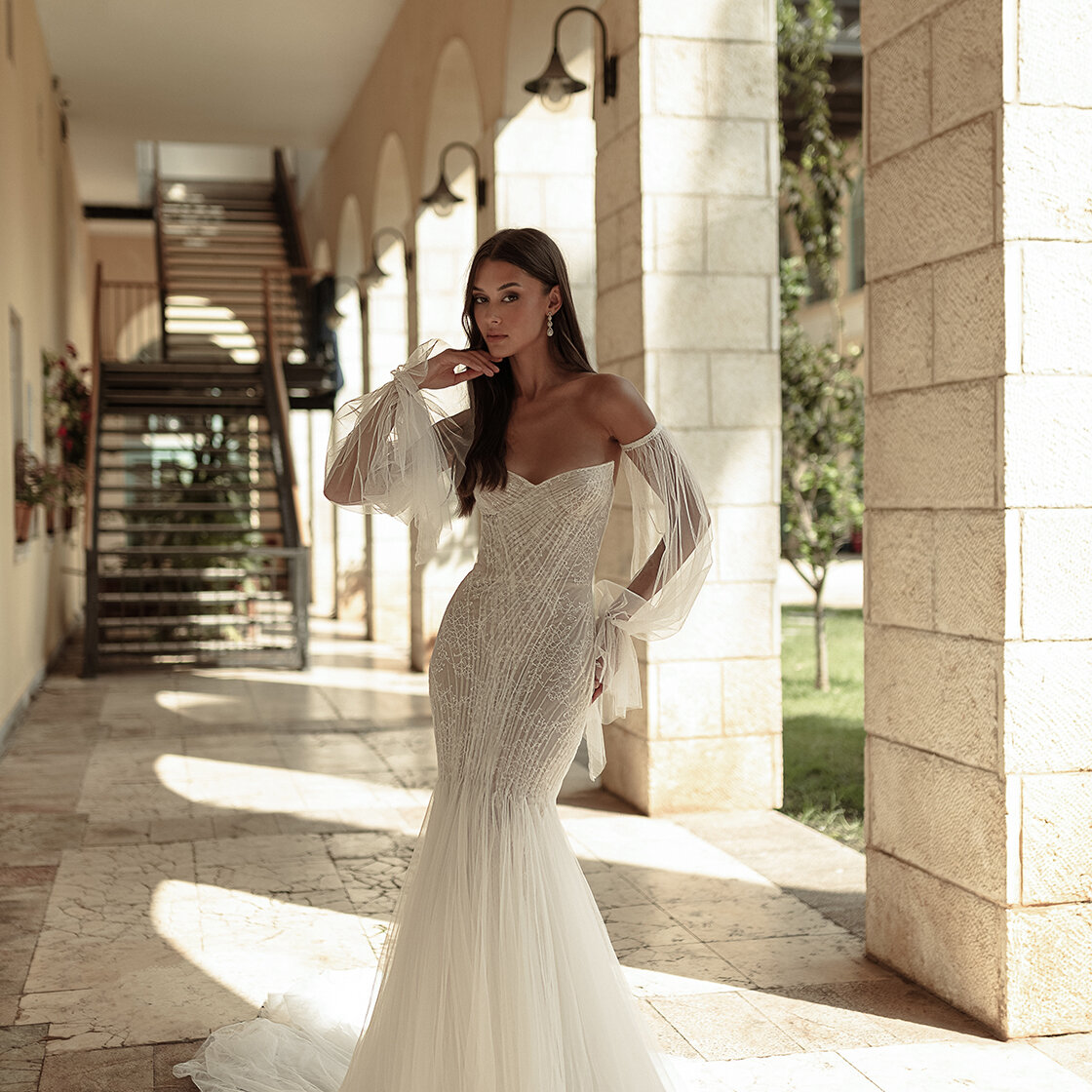 High fashion bridal portrait of a model wearing a heavily beaded, strapless mermaid gown with sheer, voluminous, detached off-the-shoulder sleeves. She stands in a sunlit Mediterranean-style stone archway.