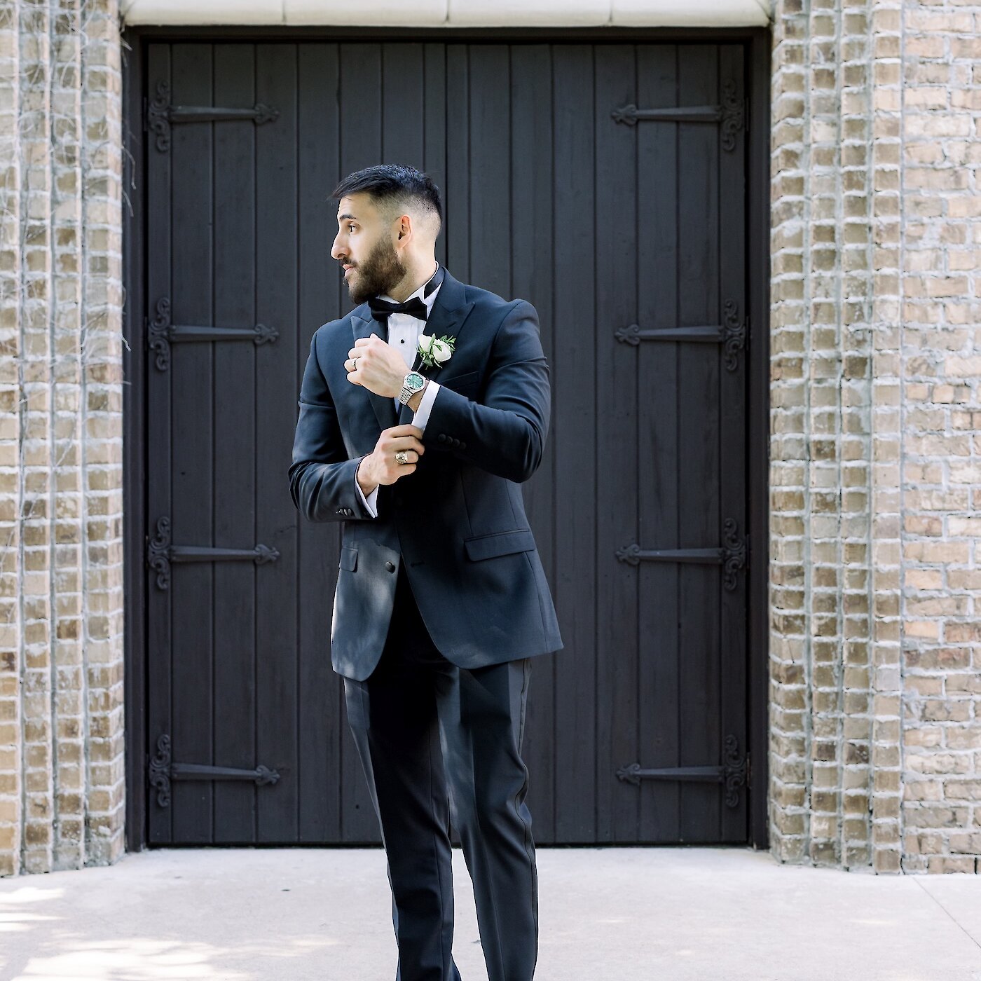 The groom, wearing a fitted black tuxedo and bow tie, adjusts his shirt cuff while posing in front of large dark wooden doors.