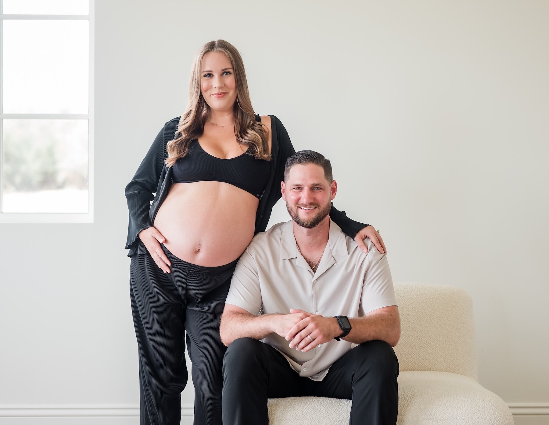 A smiling pregnant woman standing with her partner who is sitting on a white chair, posing barefoot for a bright maternity portrait.