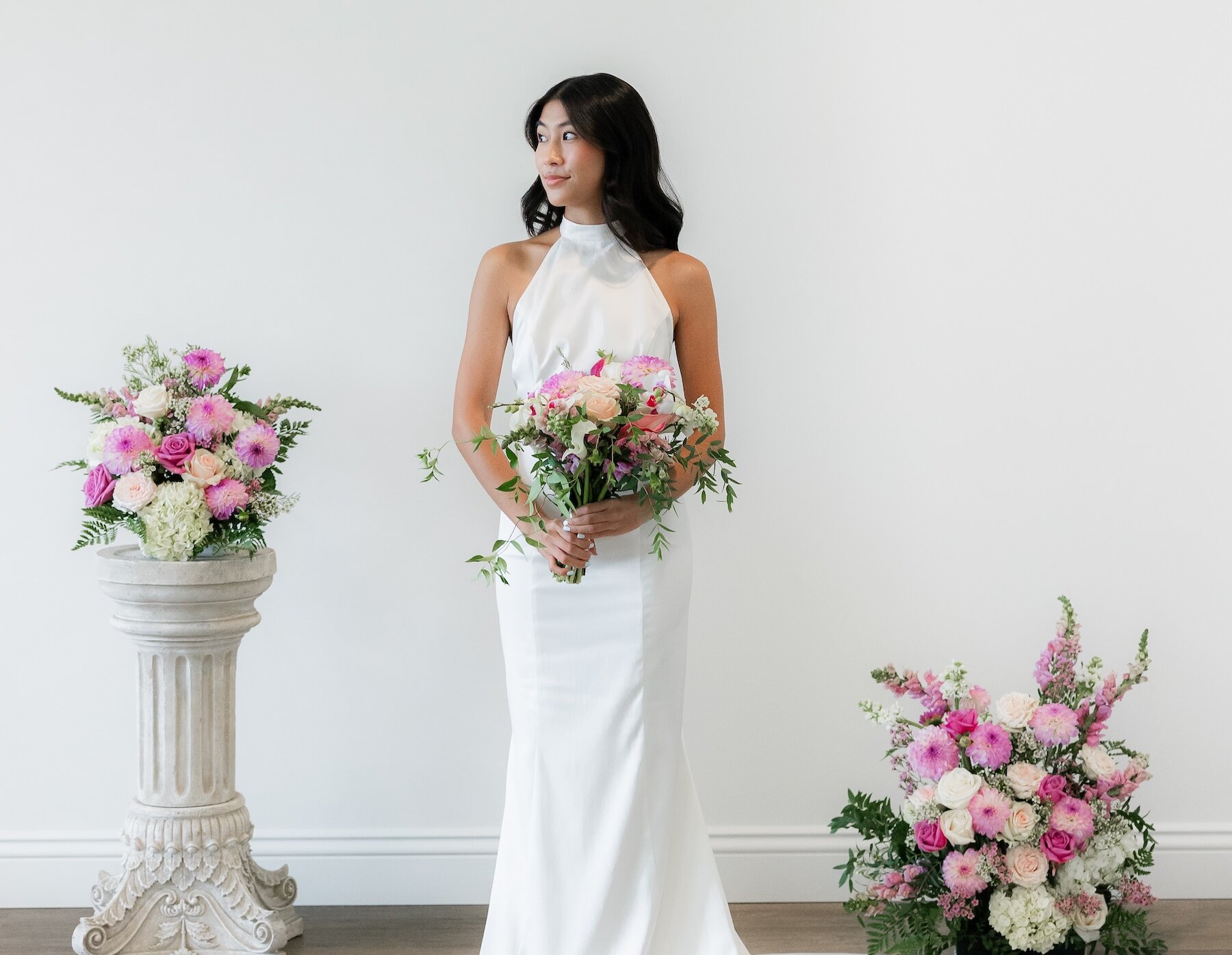 A bride wearing a sleek white halter neck gown holds a pink and white bouquet between two matching floral arrangements.