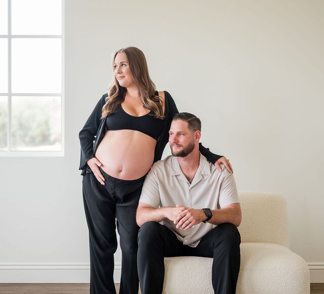 Pregnant woman standing in black loungewear with top open touching her bare stomach with her other hand on the shoulder of her male partner seated next to her