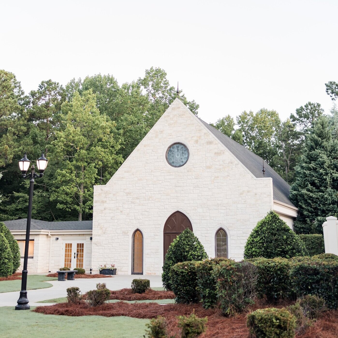 Exterior of a quaint white stone wedding chapel with an A-frame roof and arched windows, surrounded by green manicured shrubs and a forest backdrop during summer or early evening.