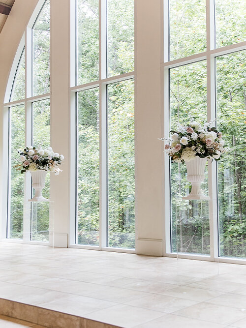 A bride from behind, wearing a strapless, white satin wedding dress with a large, dramatic bow on the back, as a woman in a navy dress zips or buttons the dress for her in a fitting room with a gold-framed mirror.