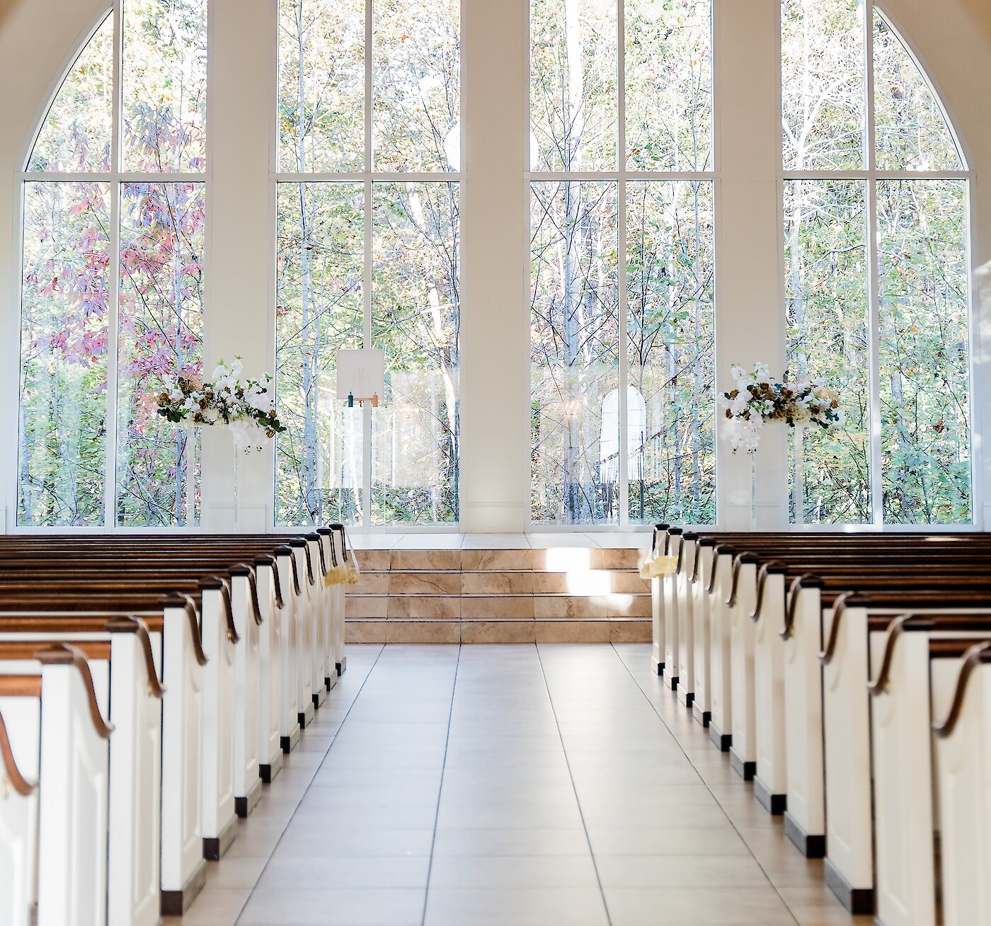 Interior of a modern chapel or wedding venue with a high A-frame ceiling and exposed wooden beams. The altar features large, tall windows overlooking an outdoor forest with fall foliage.