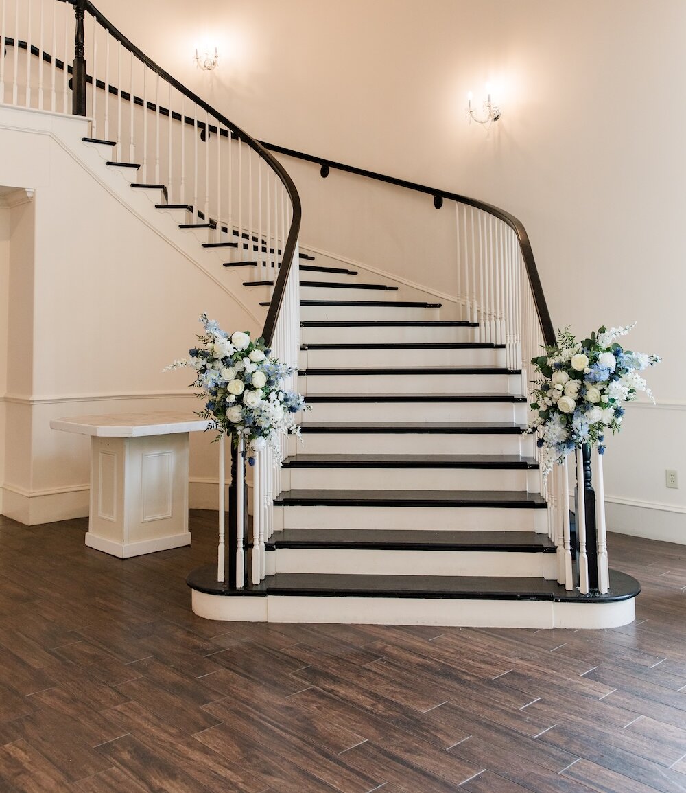 Elegant curved grand staircase with black treads, white risers, and a dark wooden handrail, decorated with large floral arrangements featuring white and blue hydrangeas and roses.