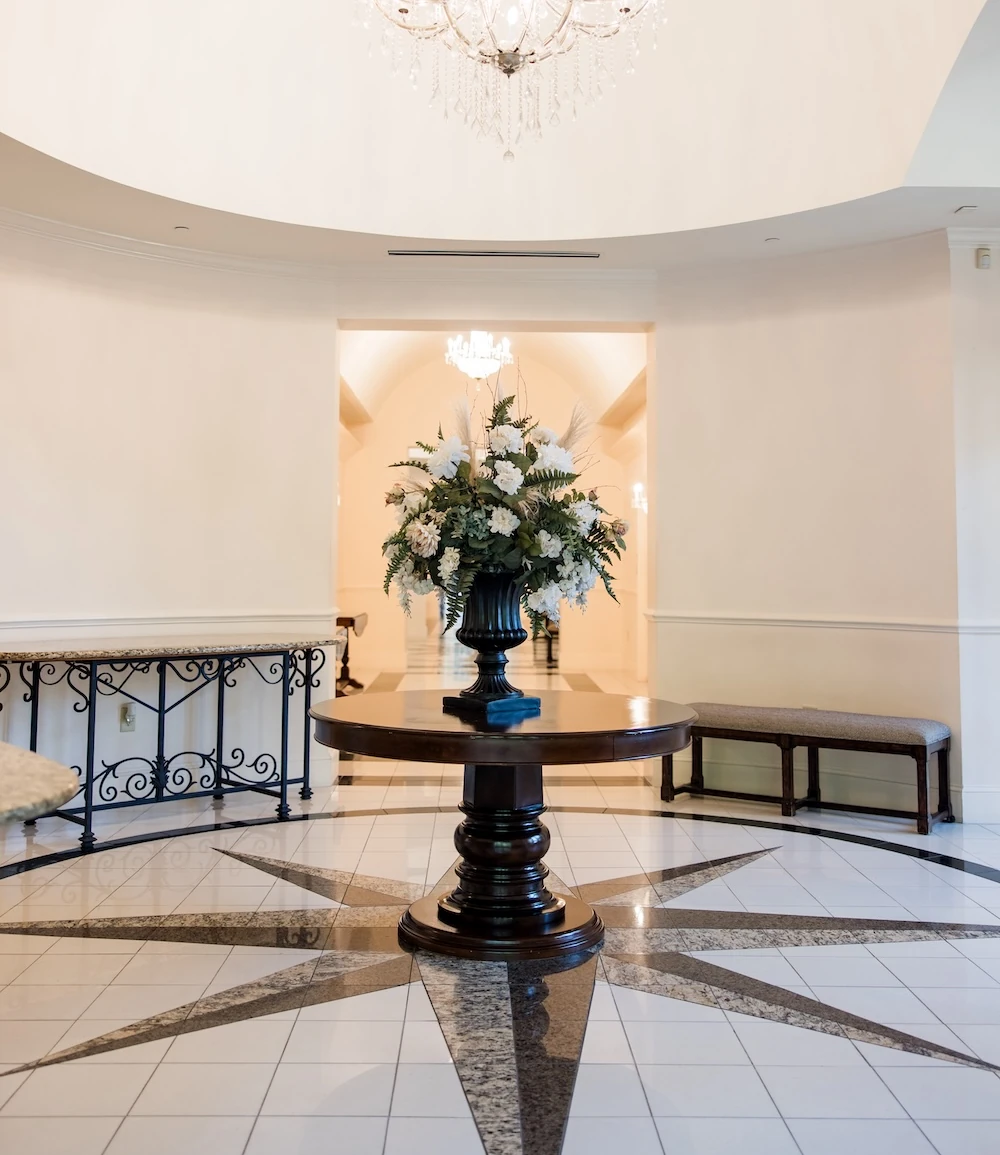 Luxury event venue foyer featuring a large crystal chandelier, a dark wood pedestal table centerpiece with a floral arrangement, and a black and white starburst pattern marble floor.