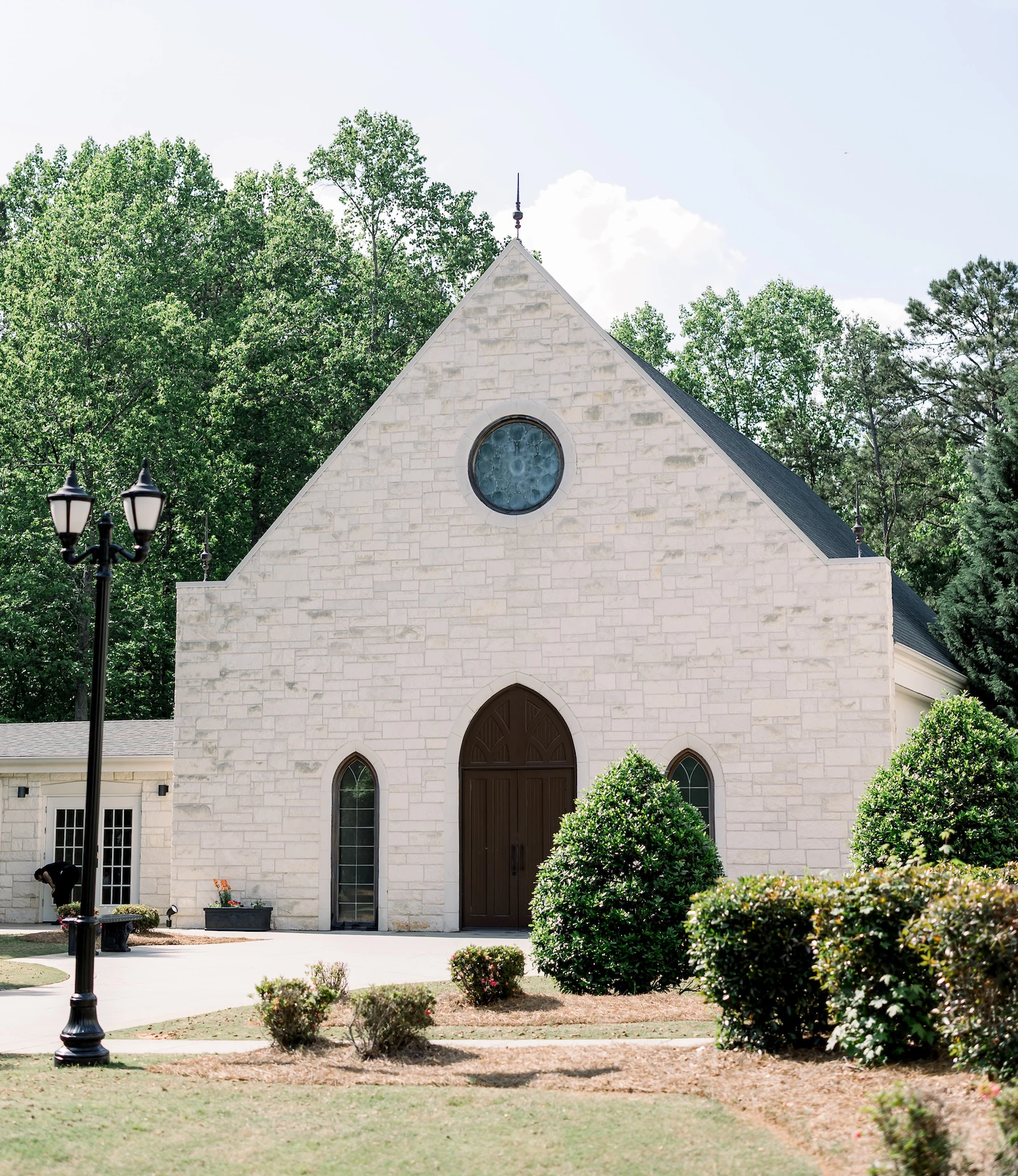 Charming stone chapel with arched wooden doors, circular stained glass window, and surrounding greenery on a sunny day.