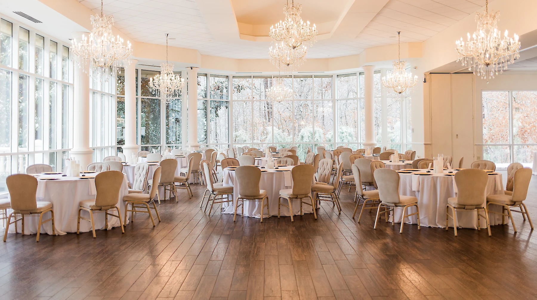 Elegant wedding reception ballroom featuring round tables set with gold chairs, multiple crystal chandeliers, and a wall of windows overlooking a snowy or wintry exterior.