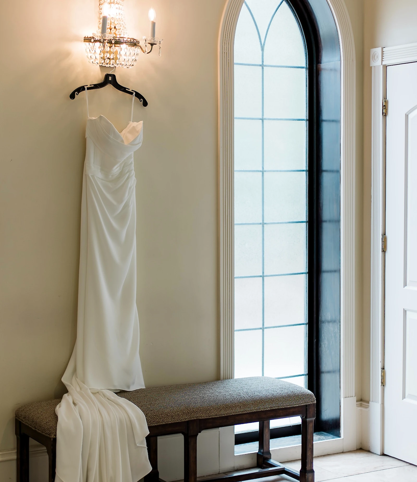 Simple, elegant white wedding dress with a sweetheart neckline and draping satin skirt, hanging on a wall next to a crystal sconce and a tall, arched Gothic-style window.