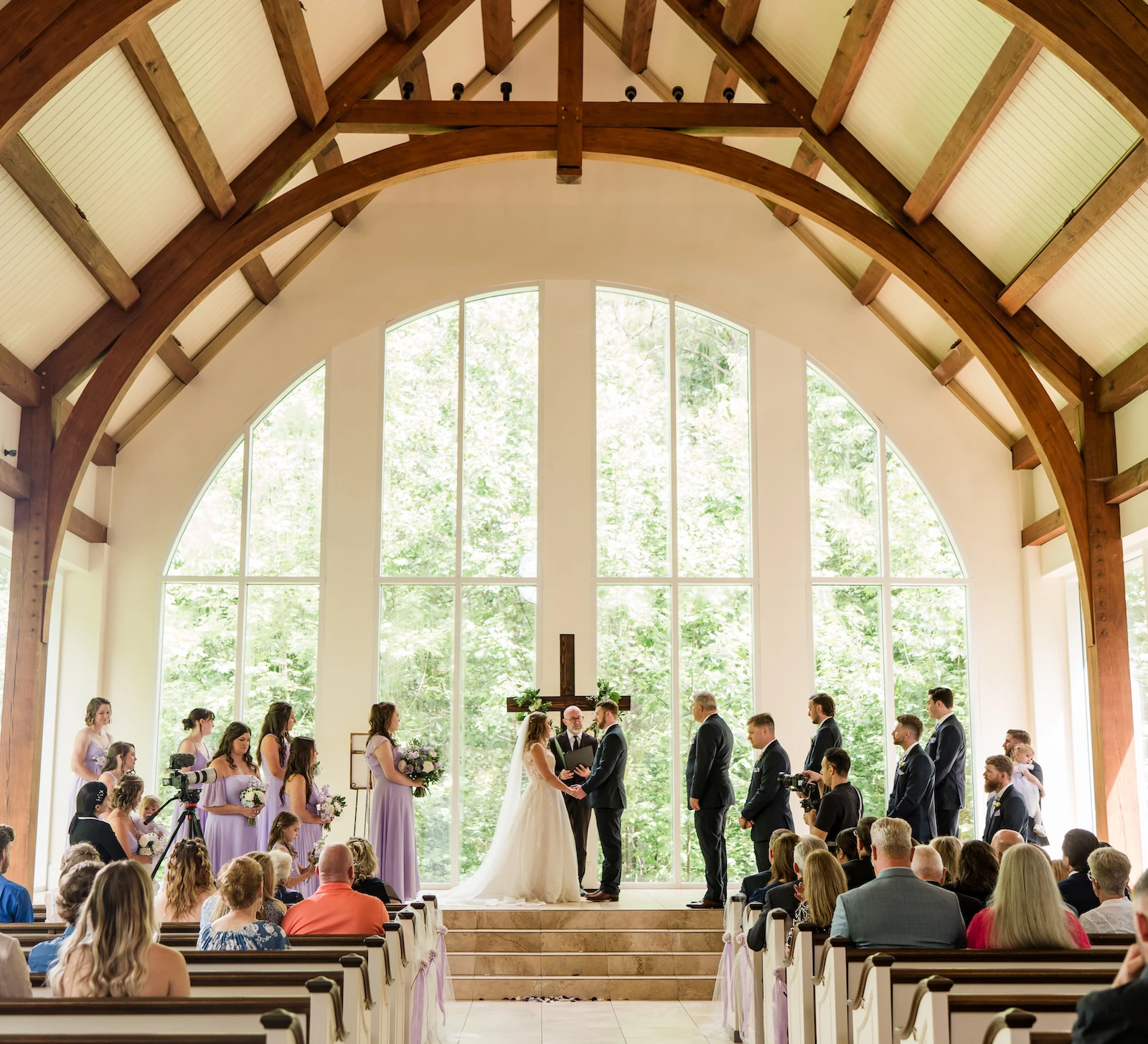 Wide view of a wedding ceremony taking place in a modern chapel with high arched wooden beams, tall windows overlooking a forest, and the wedding party standing near the altar.
