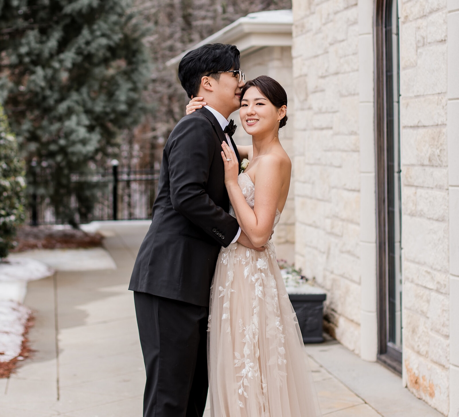 Winter wedding portrait of an Asian couple outside a stone chapel, with the groom kissing the bride's temple. The bride wears a sheer, blush-toned wedding dress with lace appliqués.