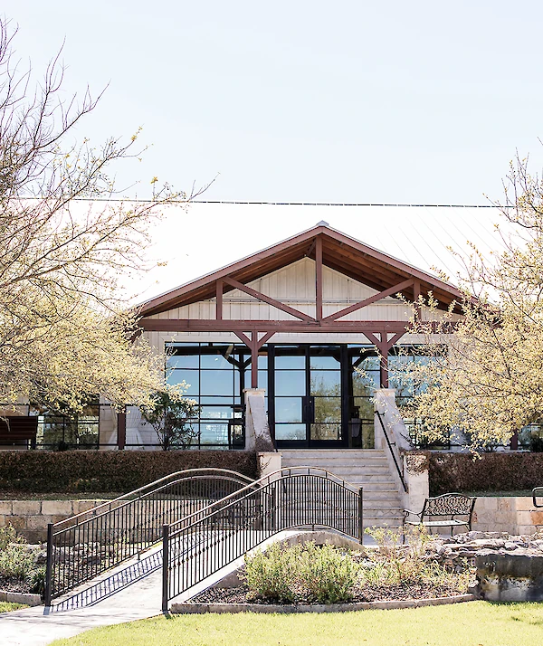 Rustic-style venue with a gabled roof, large glass windows, stone steps, and a curved metal footbridge leading to landscaped grounds.
