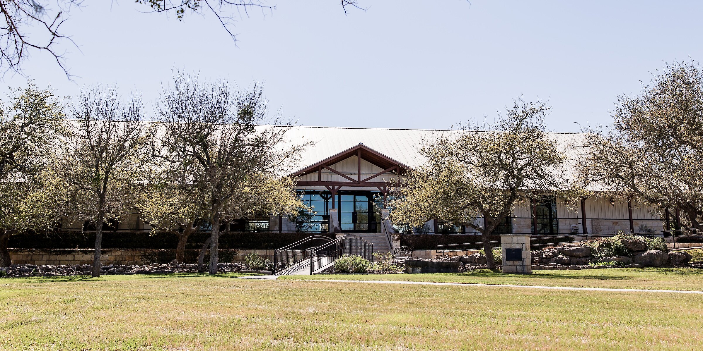 Exterior view of a rustic Texas-style wedding venue or lodge with a long, low profile, covered porch, and exposed timber beams, framed by trees and a wide grassy lawn.