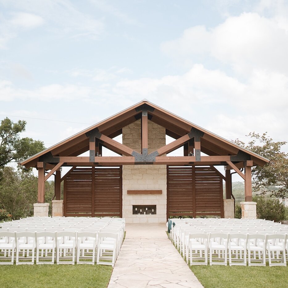 Outdoor rustic wedding ceremony setup with rows of white folding chairs facing a wooden pavilion altar with exposed beams, a stone fireplace, and a light-colored flagstone aisle.