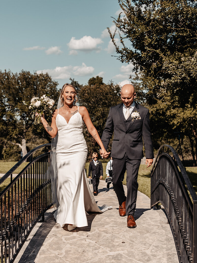 Romantic wedding portrait of a couple walking hand-in-hand through a grove of oak trees. The bride wears a lace mermaid gown and holds an orange and white bouquet, while the groom wears a black suit and rust-colored tie.
