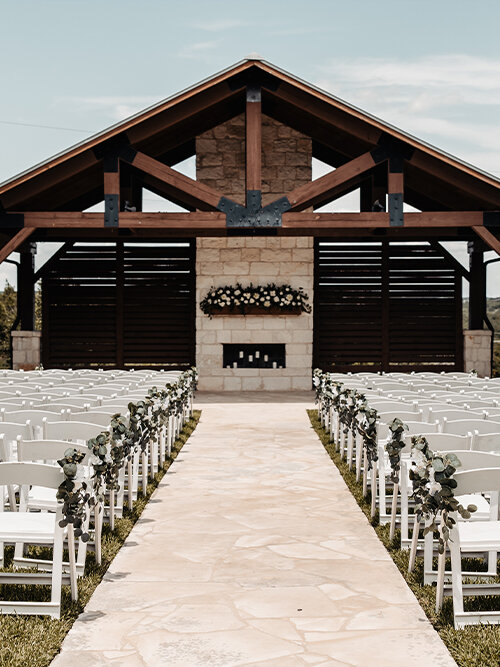 Portrait of a smiling wedding couple standing next to a white limousine under a covered rustic wooden porch. The bride holds a pastel blue and white floral bouquet.