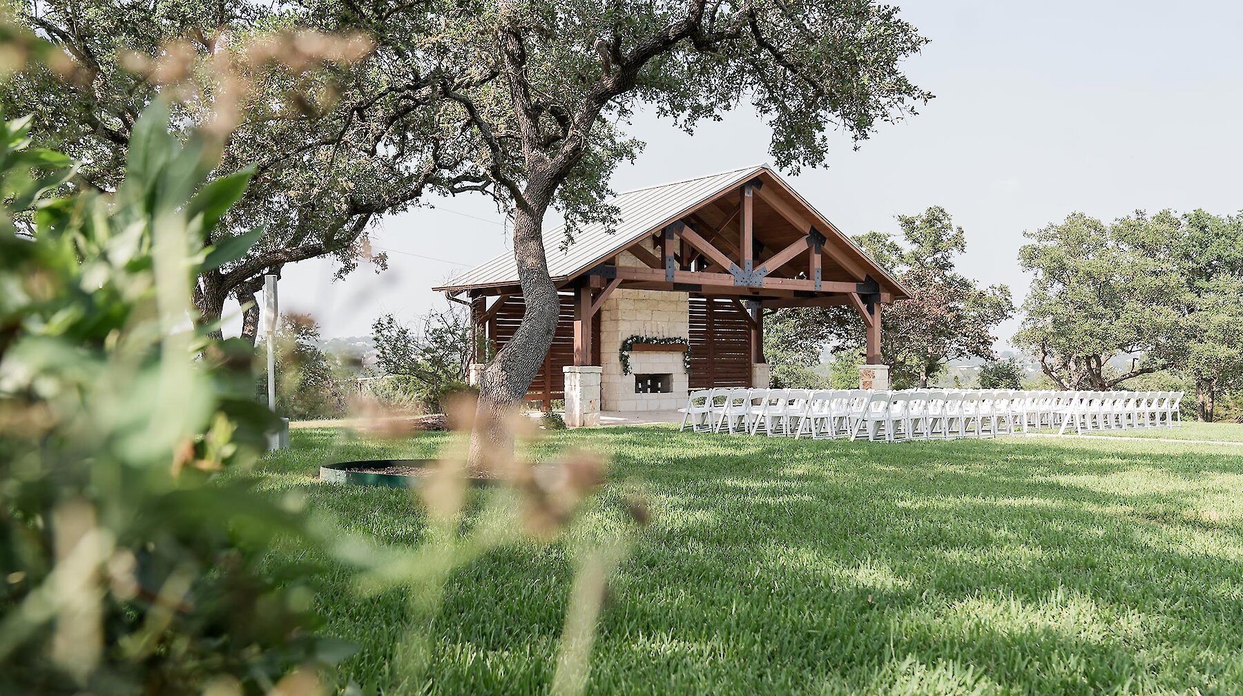 Bride and seven bridesmaids smiling in a group portrait under a wooden pergola with a stone fireplace wall decorated with a white floral garland. The bridesmaids are wearing matching sage green dresses.
