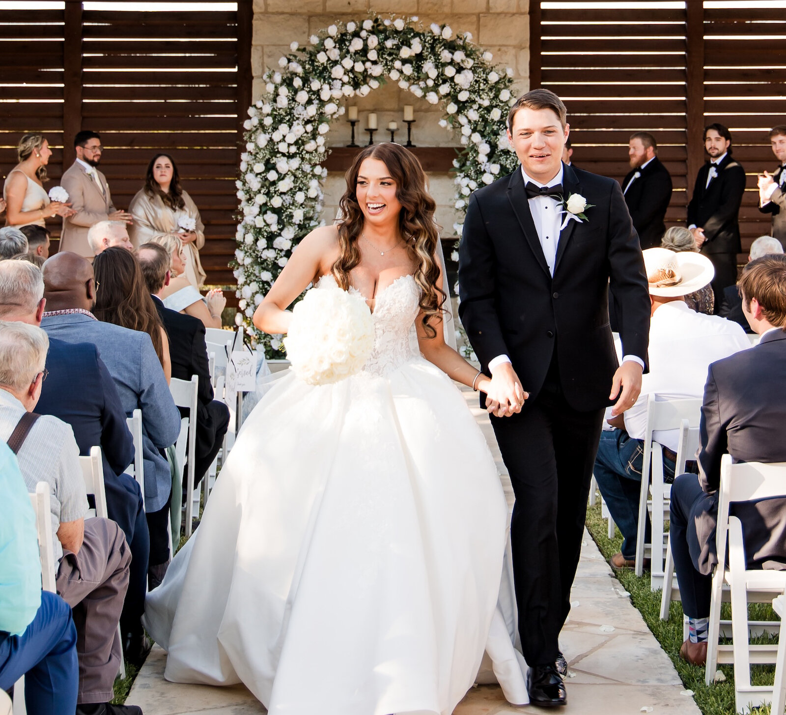 Joyful newlywed couple recessed from their ceremony at a rustic outdoor pavilion, walking down the aisle hand-in-hand past guests. The altar features a white floral arch.