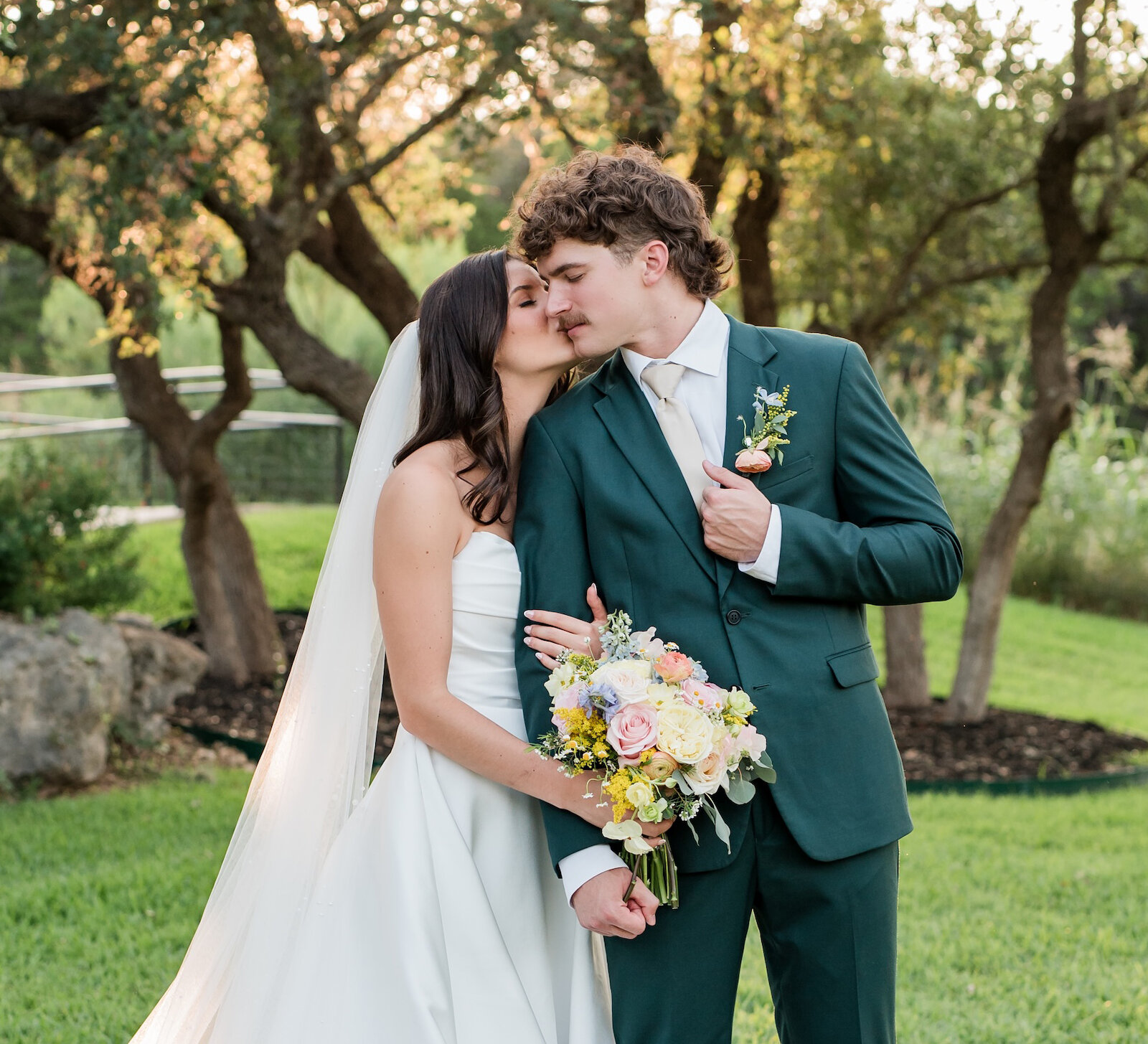 Romantic sunset wedding portrait of a couple kissing outdoors under large oak trees. The groom wears a unique dark green suit, and the bride wears a classic white satin A-line gown with a long veil.