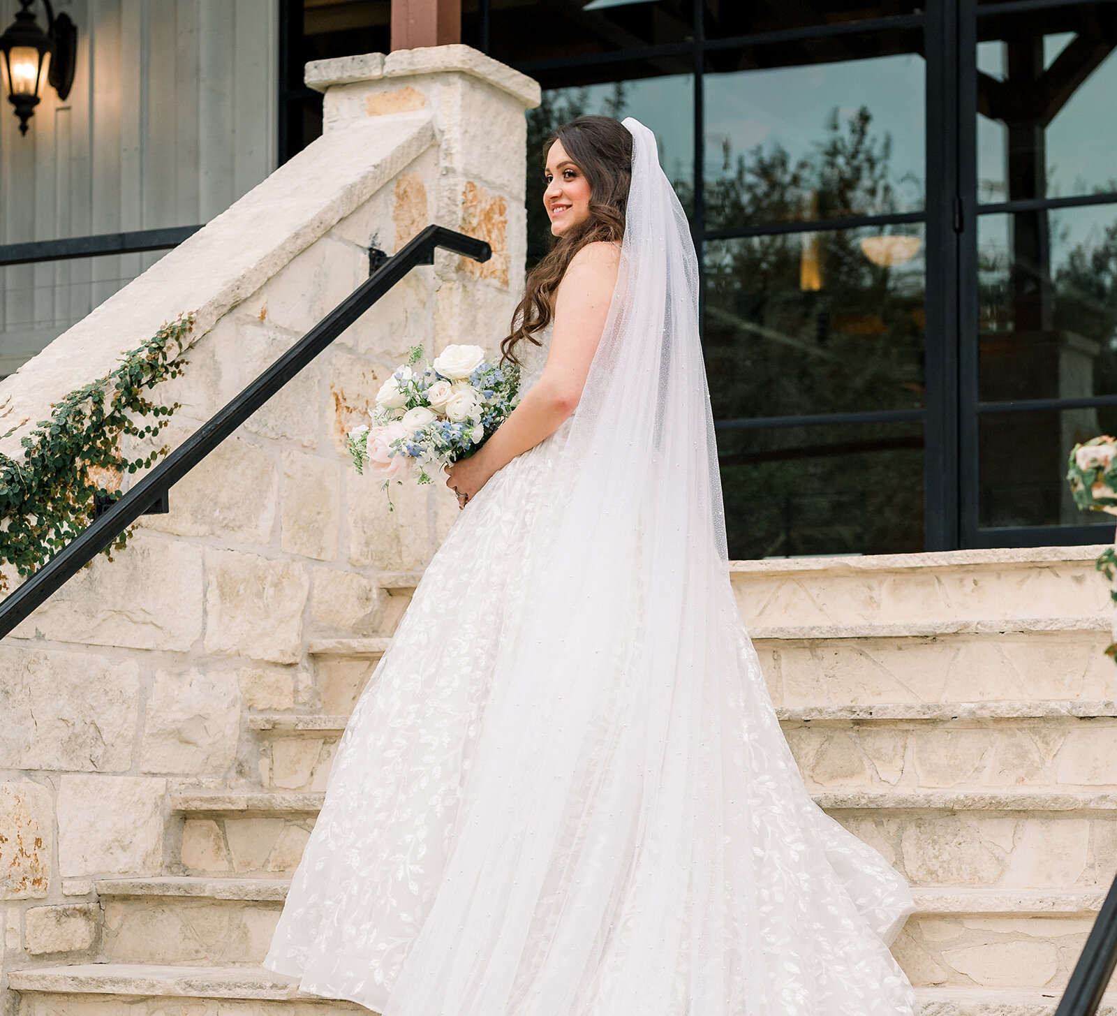 Bride in a long lace wedding gown and veil holding a white and blue bouquet while standing on stone steps outside a venue
