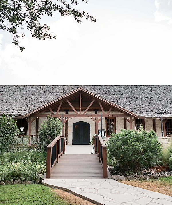 Rustic stone venue with dark wooden trusses, double arched doors, and a wooden footbridge leading through landscaped greenery
