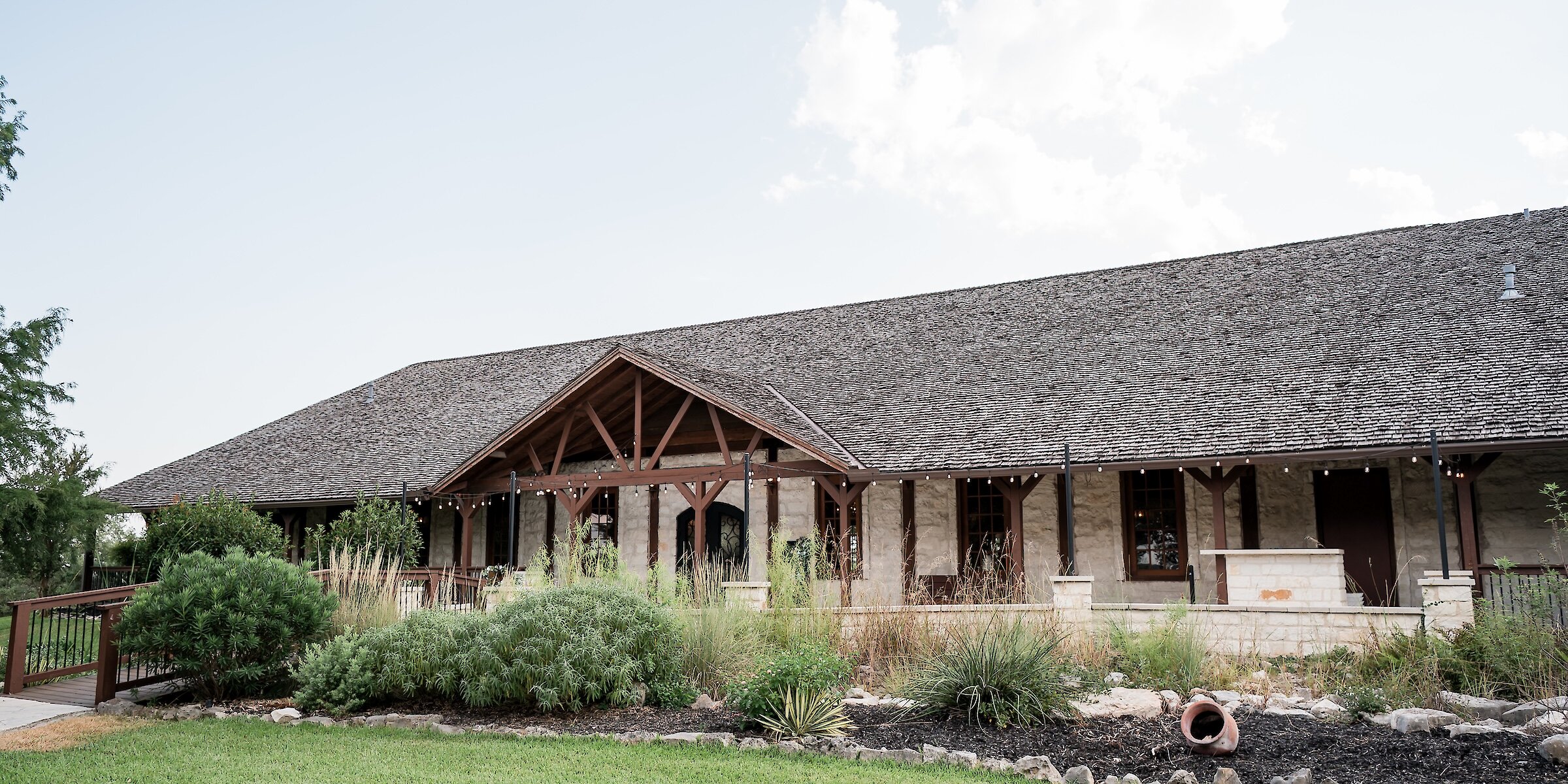 Exterior of a sprawling rustic wedding venue with a low-slung shingled roof, light stone facade, exposed wooden beams on the covered entryway, and native landscaping in the foreground.