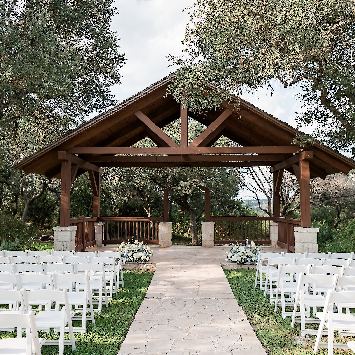 Outdoor wedding ceremony setup featuring a wooden pavilion altar with exposed beams, rows of white folding chairs, a flagstone aisle, and floral arrangements of white and blue hydrangeas, all set beneath large oak trees.