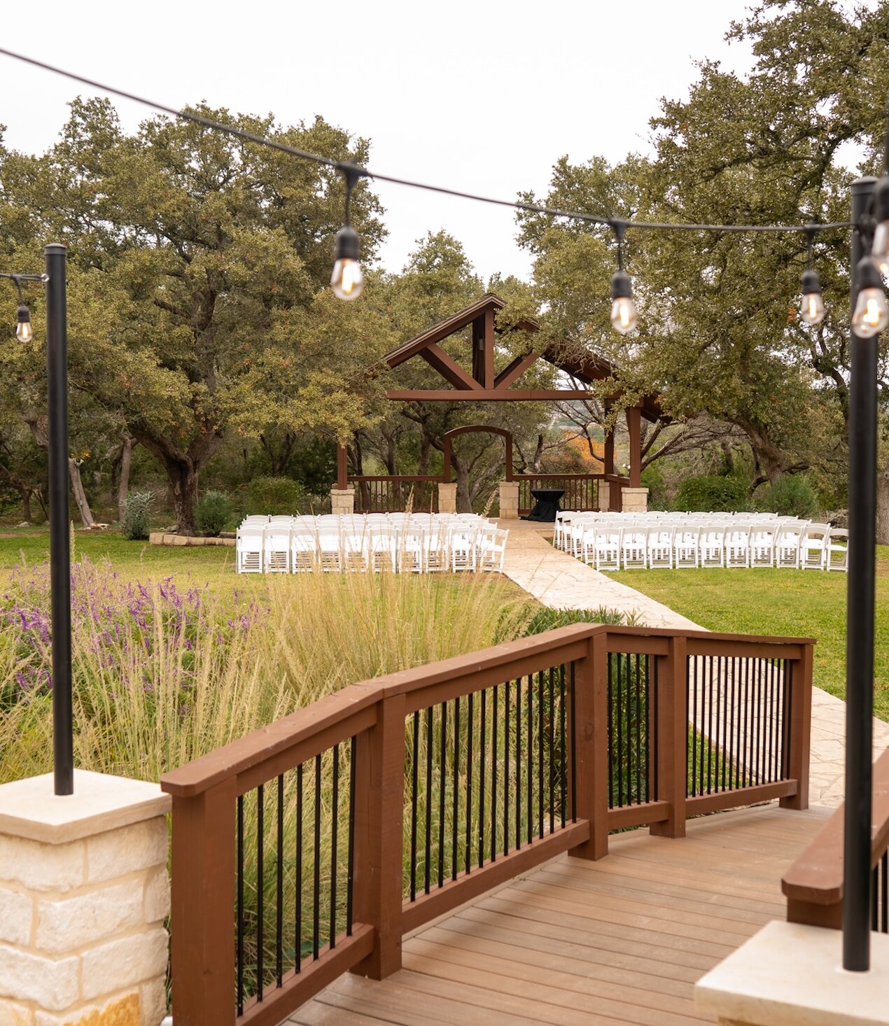 Outdoor rustic wedding ceremony setup with white chairs on a green lawn leading to a wooden pavilion altar, viewed over a wooden deck with string Edison lights overhead.