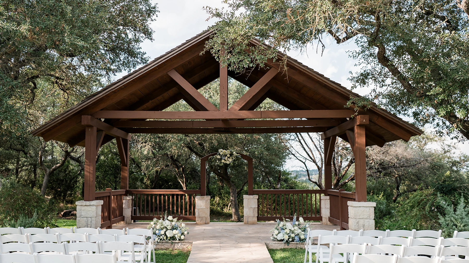 Outdoor wedding ceremony pavilion with wooden beams, stone columns, white chairs, and floral arrangements surrounded by trees