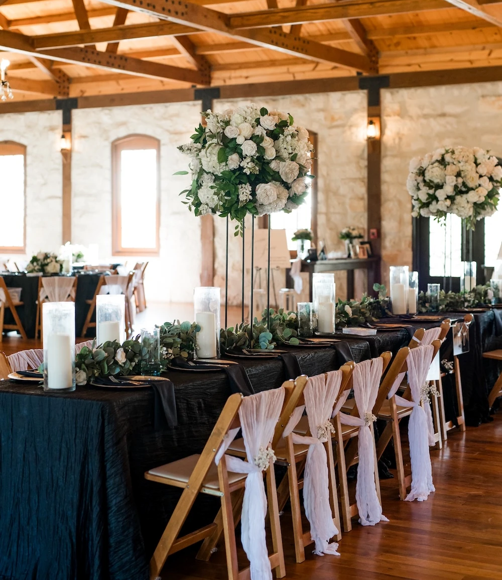 Elegant wedding reception head table featuring black crinkled linens, tall white floral centerpieces, eucalyptus garlands, and gold Chiavari chairs decorated with sheer blush fabric sashes in a rustic wooden beamed venue.