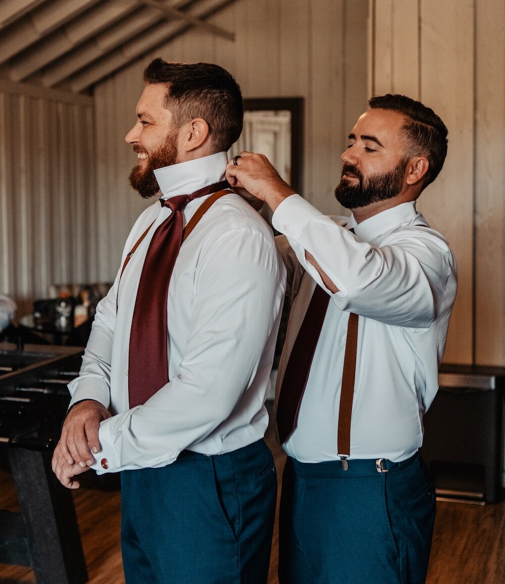 Groom getting ready for the wedding, standing in a rustic, wood-beamed suite while a groomsman helps him with his burgundy tie and shirt collar. Both men wear suspenders and navy blue pants.
