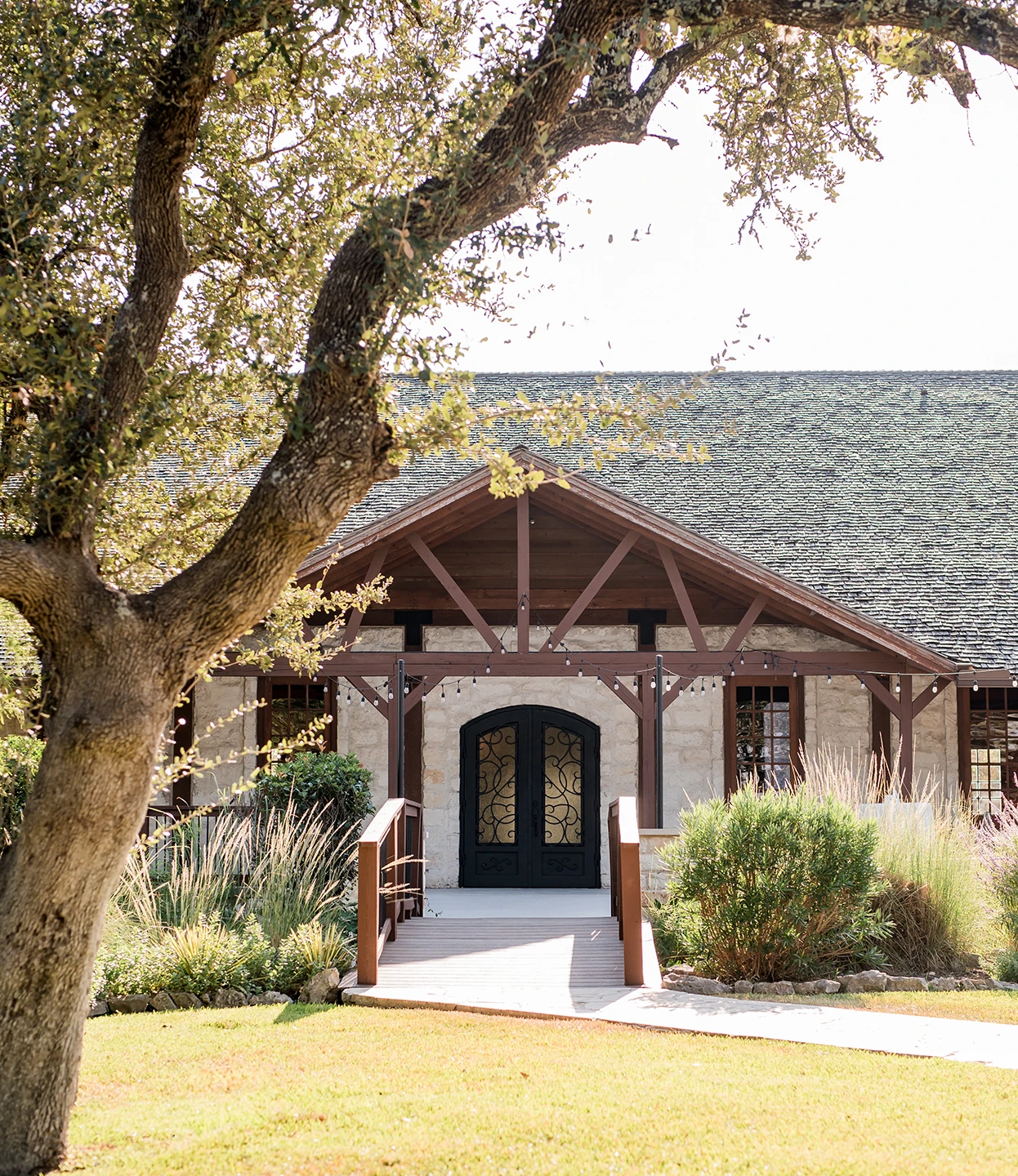 Rustic stone wedding venue with wooden beams, arched double doors, and a tree framing the entrance on a sunny day