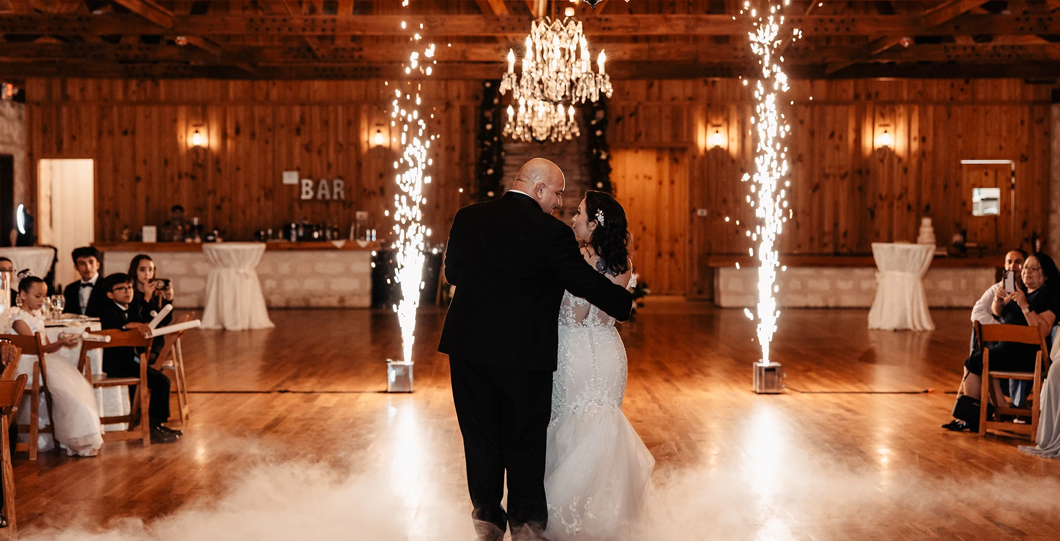 Romantic wedding first dance in a rustic reception hall with wood-plank walls and exposed beams. The couple dances on a cloud of fog, flanked by two cold sparkler fountains, beneath crystal chandeliers.