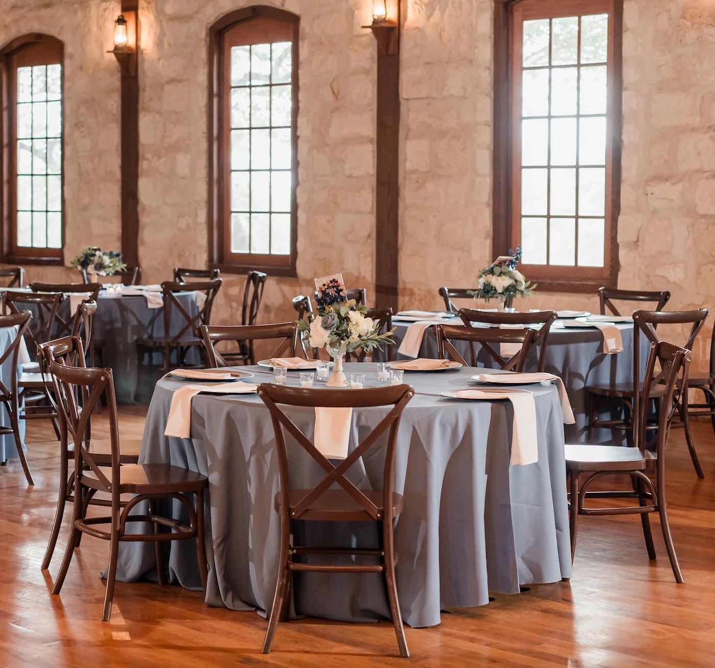 Rustic wedding reception hall with stone walls, wooden cross-back chairs, blue table linens, and floral centerpieces