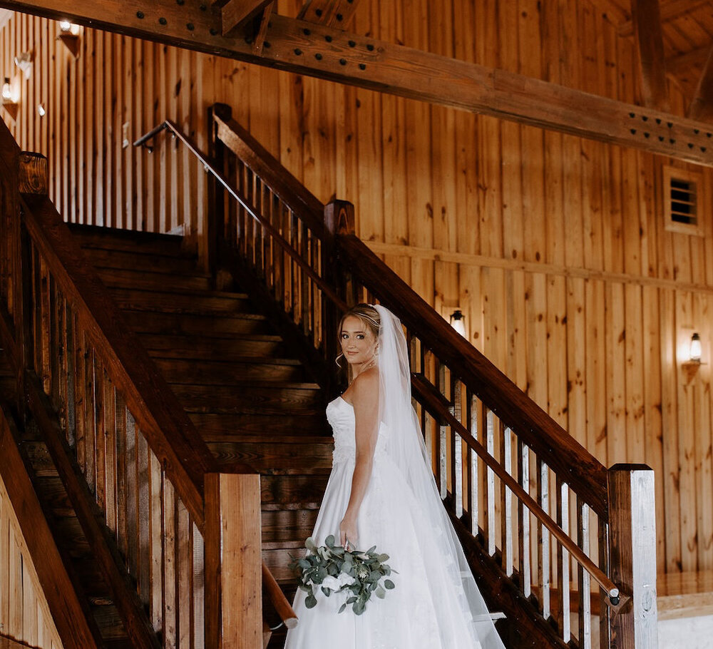 Bridal portrait of the bride descending a rustic wooden staircase in a barn wedding venue. She wears a strapless white gown with a long veil and holds a bouquet of white florals and eucalyptus greenery.