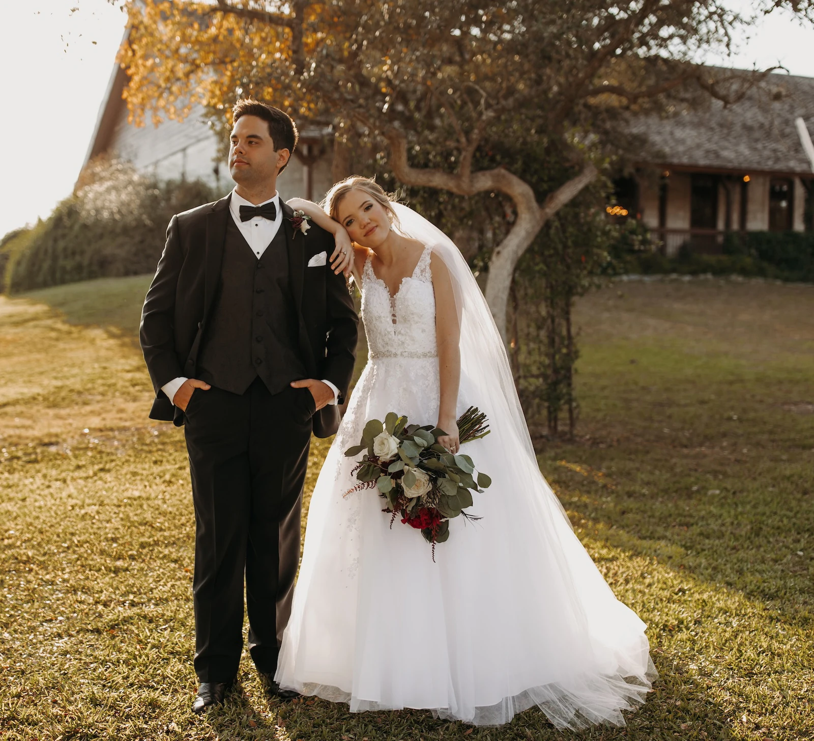 Romantic golden hour wedding portrait of the bride and groom standing outdoors near a large tree. The groom wears a black tuxedo, and the bride leans on his shoulder, wearing a lace gown and holding a burgundy and eucalyptus bouquet.