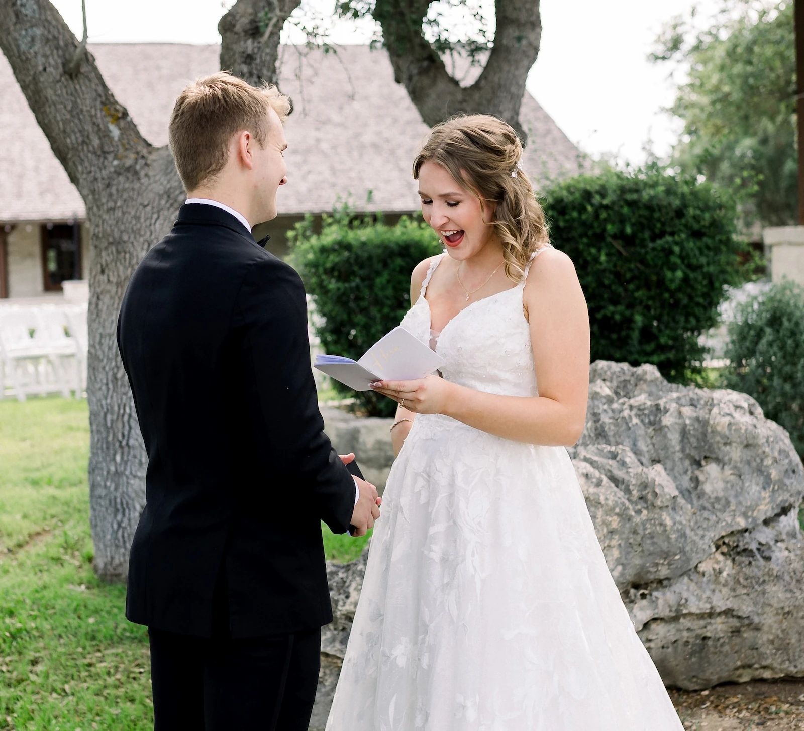 Emotional outdoor wedding photo showing the bride laughing and reacting to the groom while reading handwritten vows under a large tree. The background includes a venue building and outdoor seating.