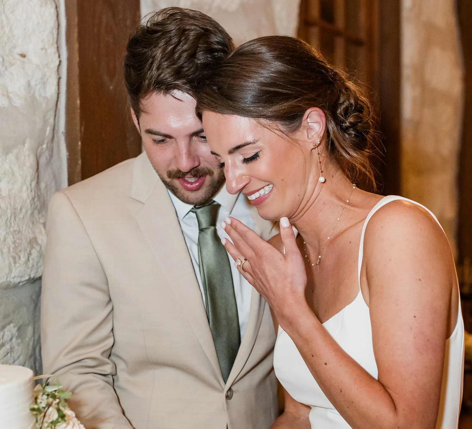 Bride and groom laughing while cutting their three-tiered wedding cake with white and blush floral accents. The groom wears a tan suit and the bride wears a simple white slip dress, standing against a rustic stone wall.
