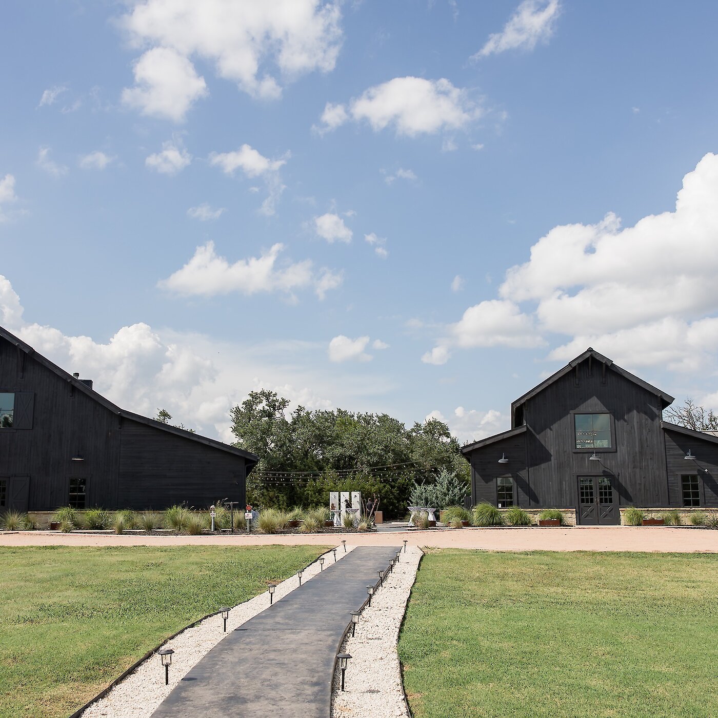 Wide exterior view of a modern black barn wedding venue complex under a blue, cloudy sky. Two black barn-style buildings flank a grassy lawn with a central gray walkway and landscape lighting.