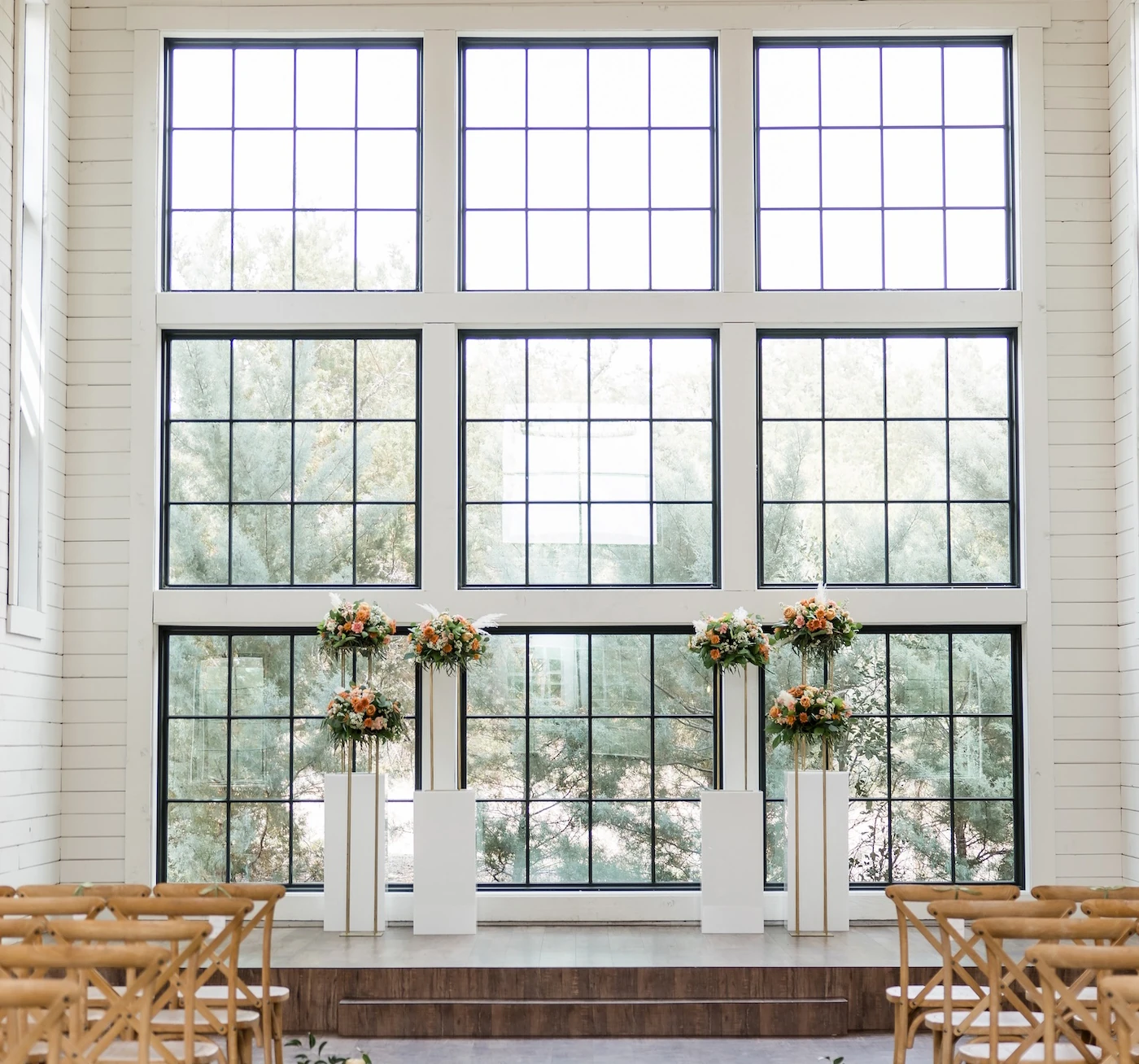Bright indoor wedding ceremony setup with large grid windows, floral arrangements on modern white stands, and wooden cross-back chairs