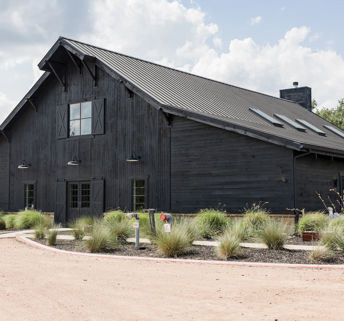 Modern black barn-style event venue with metal roof, front landscaping, and cloudy sky background
