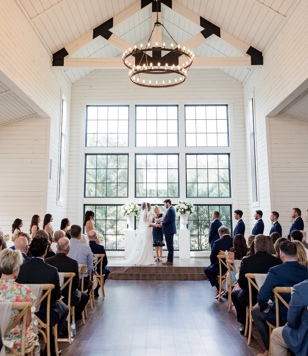 Modern farmhouse wedding ceremony interior with a vaulted white ceiling and exposed trusses, featuring a couple at the altar in front of large black-grid windows. Guests are seated on cross-back wooden chairs under large wagon wheel chandeliers.