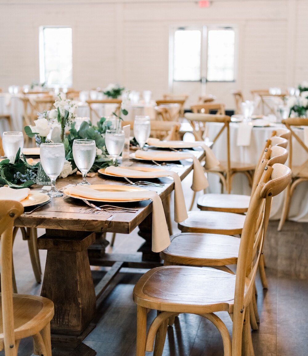 Rustic wedding reception table set with gold charger plates, light wooden chairs, and a floral garland of white roses and eucalyptus on a long, dark wood farm table inside a bright, white-walled venue.