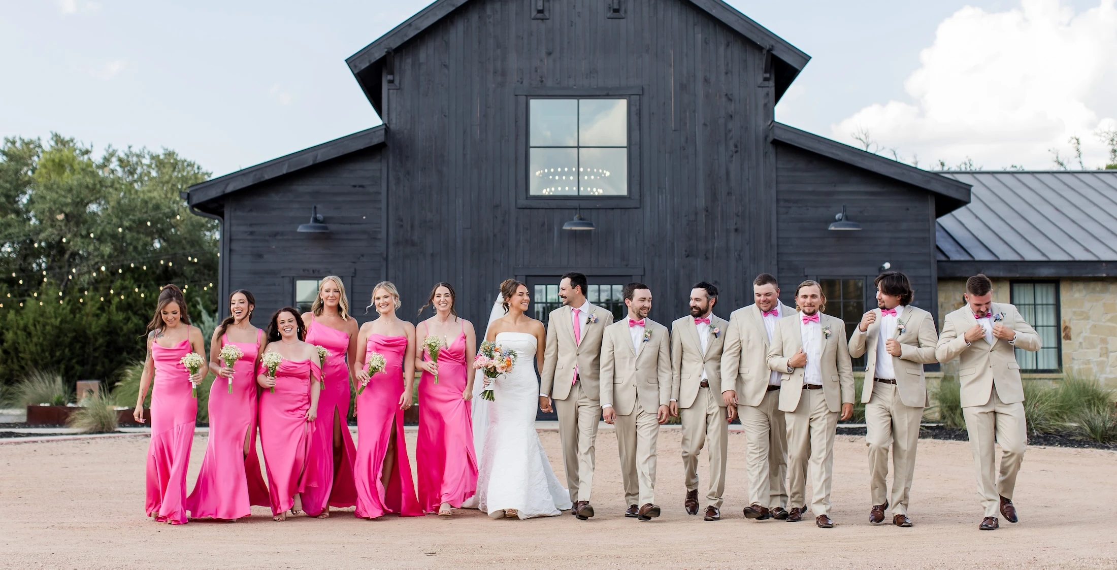 Large, vibrant wedding party walking and smiling in front of a modern black barn venue. Bridesmaids wear long hot pink satin dresses, and groomsmen wear tan suits with pink ties and bow ties.